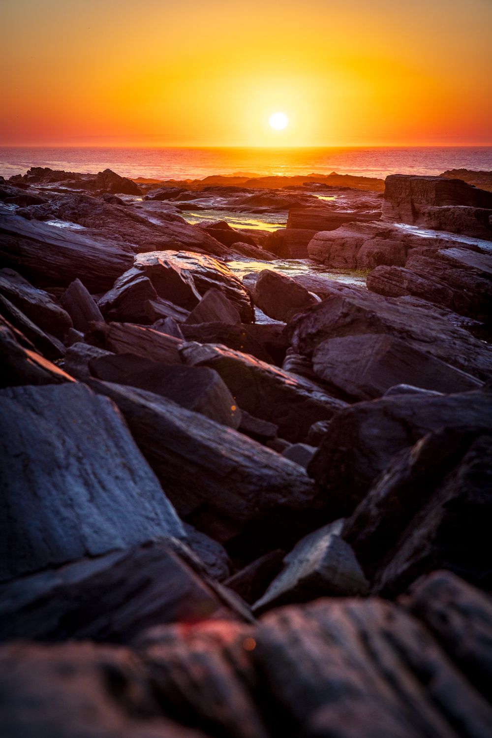 Rocky coastline at sunset with vibrant orange sky and sun setting over the ocean horizon.