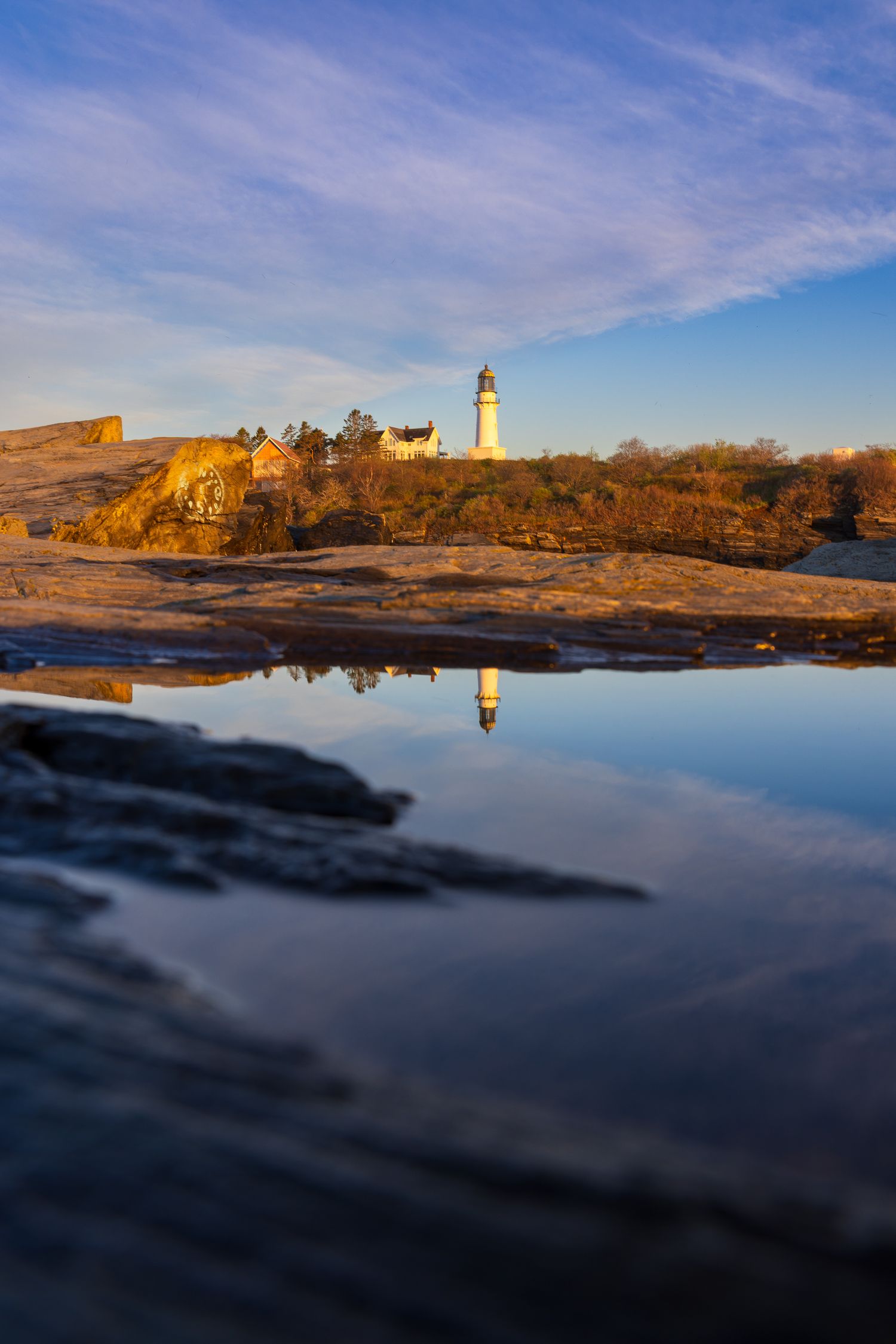 A lighthouse reflects in a tidal pool on rocky New England coast at golden hour under blue skies.