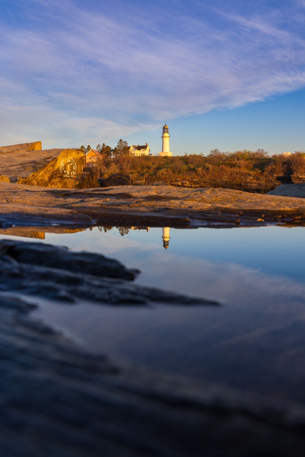 A lighthouse reflects in a tidal pool on rocky New England coast at golden hour under blue skies.