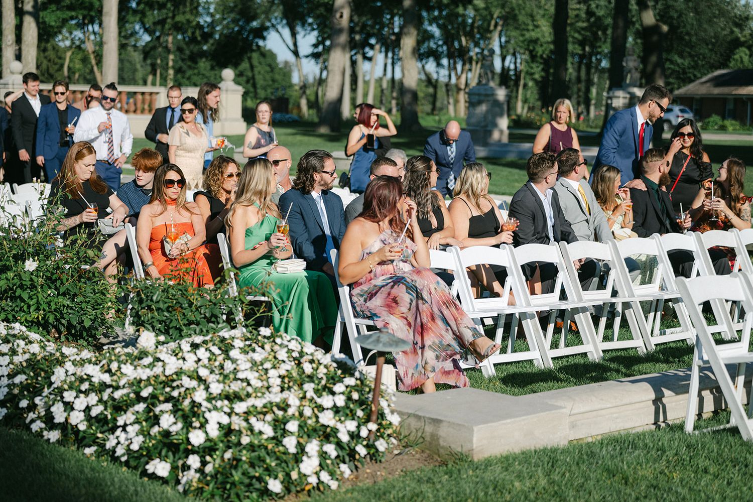 Armour House Wedding - Guests seated in white chairs at an elegant outdoor wedding ceremony on a sunny day with lush green landscaping.