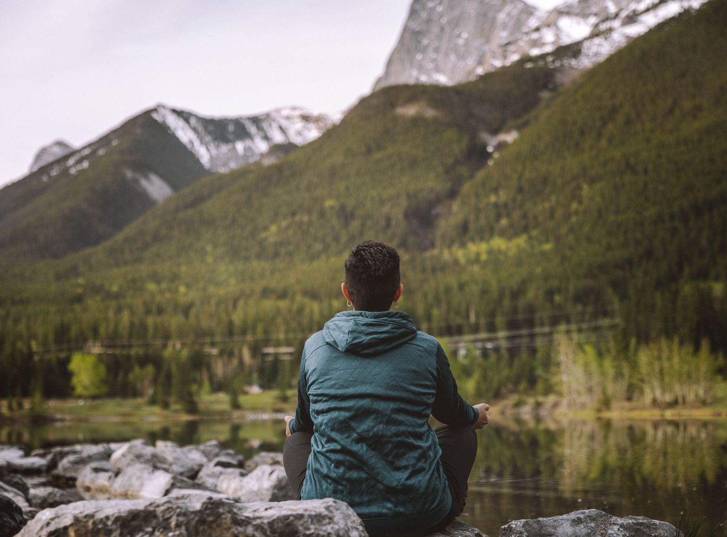 A man sits cross-legged on rocks by a calm mountain lake, gazing at snow-capped peaks reflected in the still water.