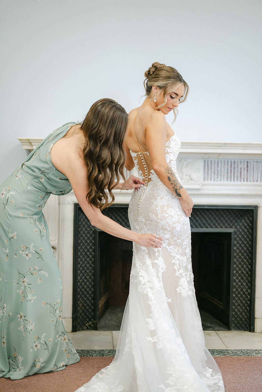 Armour House Wedding - Bridesmaid in sage green floral gown buttoning the back of a bride's white lace wedding dress near a fireplace.