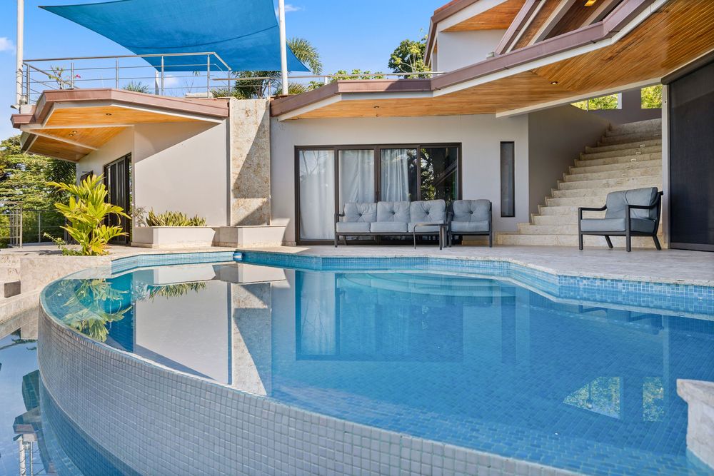Luxury villa with a curved infinity pool, glass railings, and modern outdoor seating area surrounded by lush tropical landscaping.