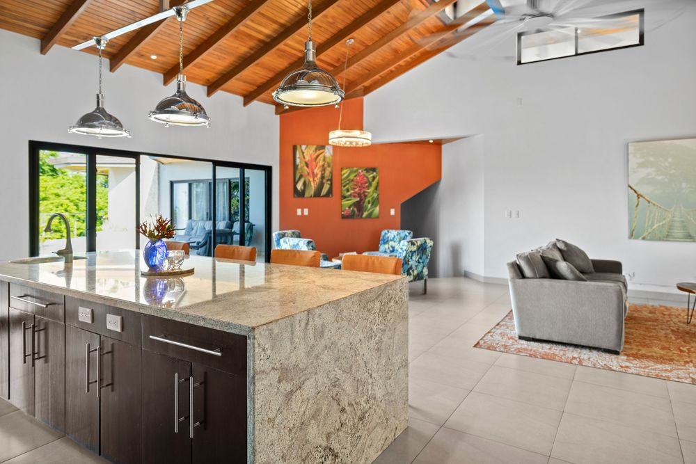 Modern open-plan kitchen with granite island, vaulted wood ceiling, pendant lights, and living area with orange accent wall.