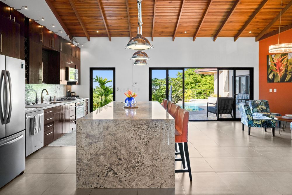 Modern kitchen featuring a large granite island, stainless steel appliances, wood beam ceiling, and open view to tropical pool area.