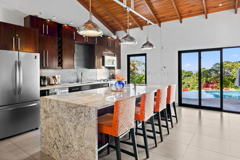 Modern kitchen with granite island, orange bar stools, dark cabinets, stainless appliances, vaulted wood ceiling, and pool view.