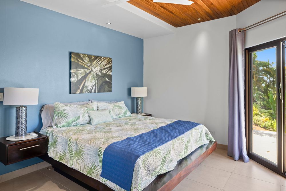 Tropical bedroom with blue accent wall, wood ceiling, king bed with floral bedding, and sliding glass door to garden.