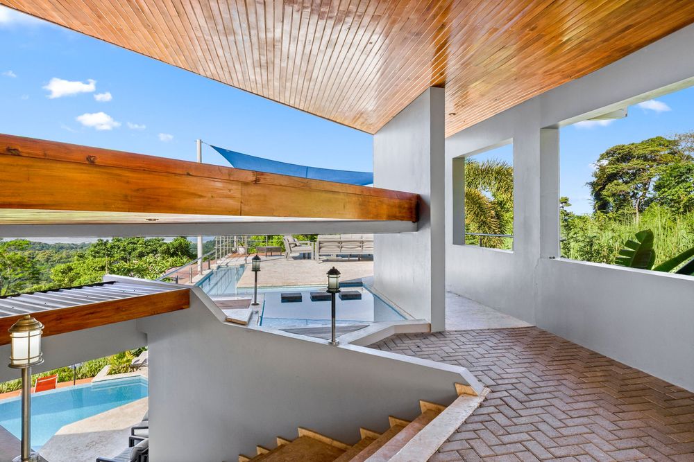 Spacious covered balcony with wood ceiling, white railings, curved staircase, and scenic tropical pool view below.