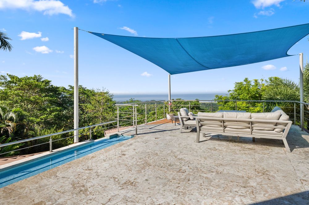 Elevated terrace with blue shade sail, outdoor sofa, pool edge, and panoramic tropical ocean views.