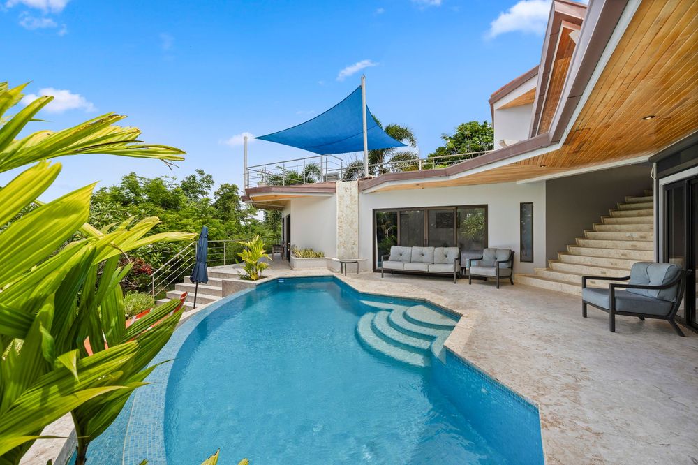 Luxury villa with curved pool, blue shade sail, tropical landscaping, and spacious stone patio with outdoor seating.