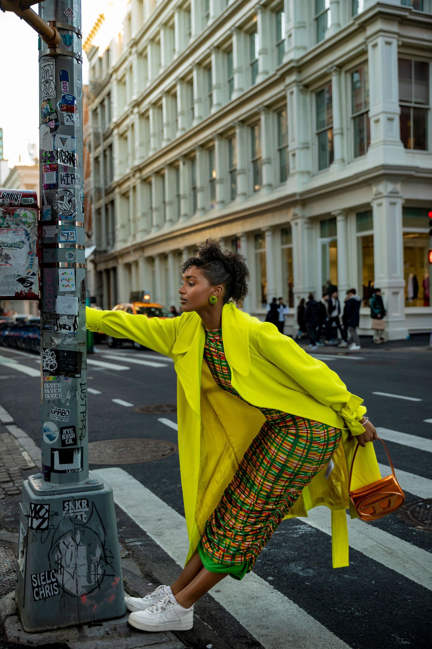 Woman in neon yellow coat and plaid dress poses on NYC crosswalk, leaning on sticker-covered pole.
