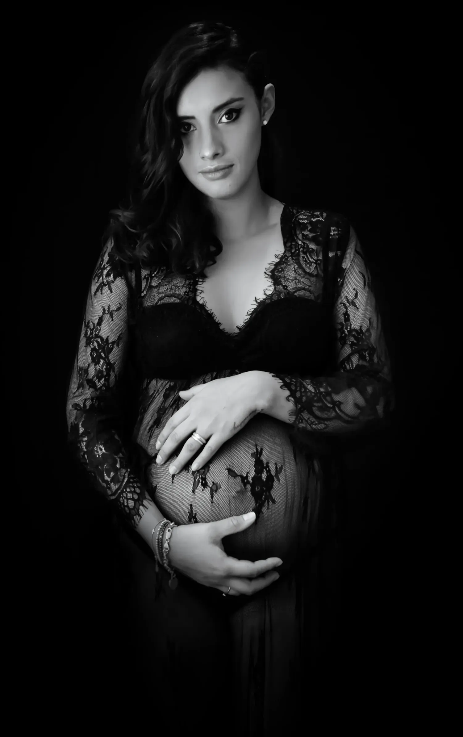 Photo de maternité en noir et blanc d'une femme enceinte en robe de dentelle noire, caressant son ventre rond sur fond sombre.