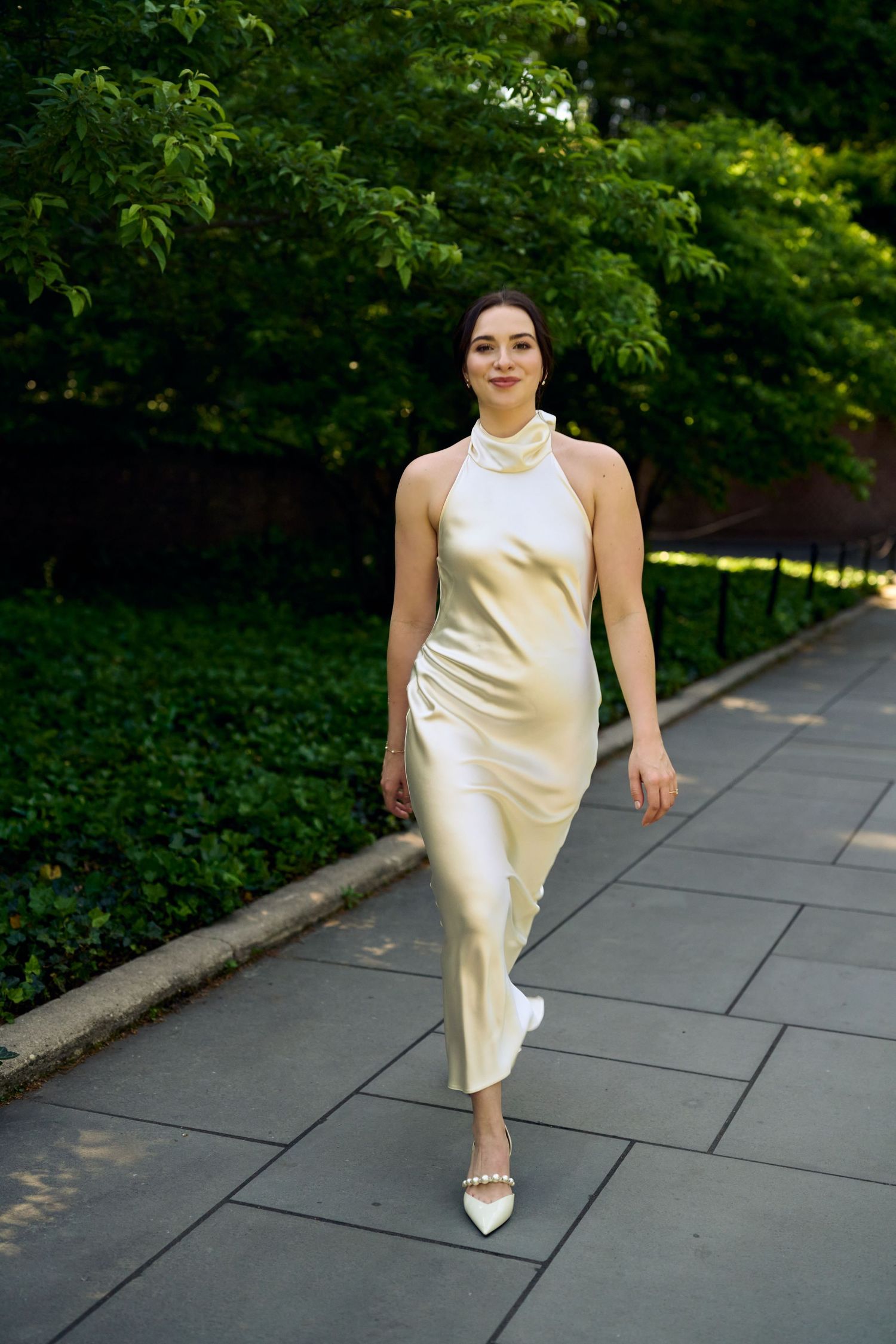 Woman in elegant ivory satin halter midi dress and white heels walking on a garden path surrounded by lush green trees.