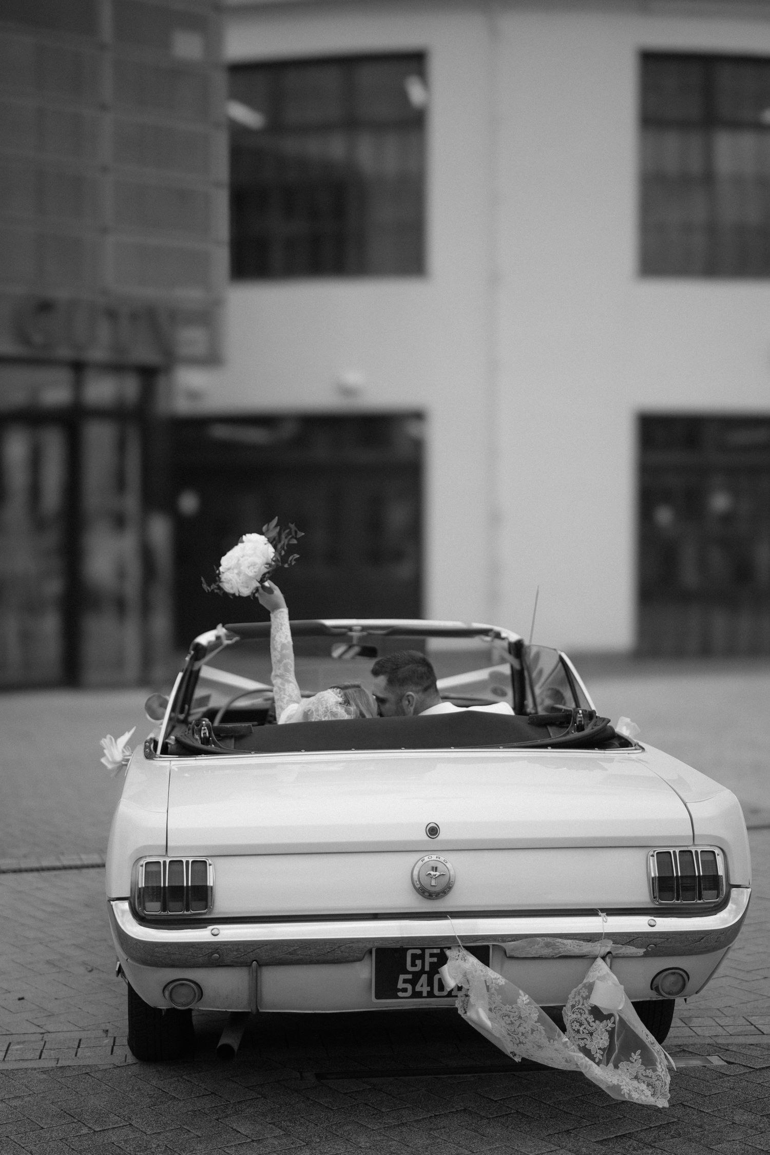 Black and white photo of a classic Ford Mustang convertible decorated for a wedding, with a bouquet visible.