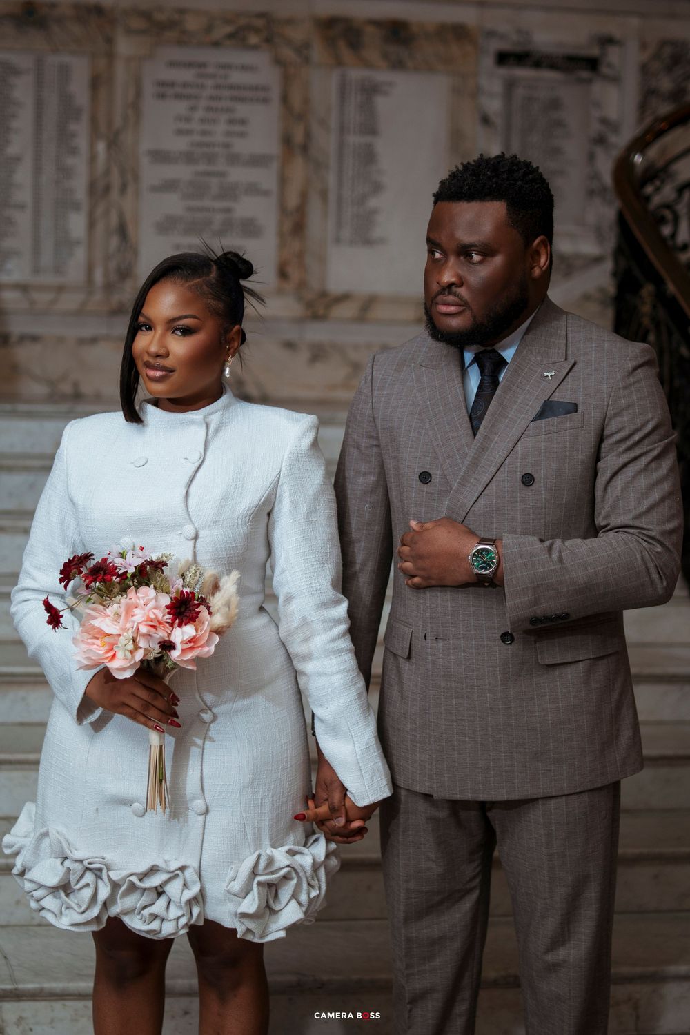 Elegant couple in wedding attire, bride in white dress with floral hem holding bouquet, groom in gray suit holding hands.