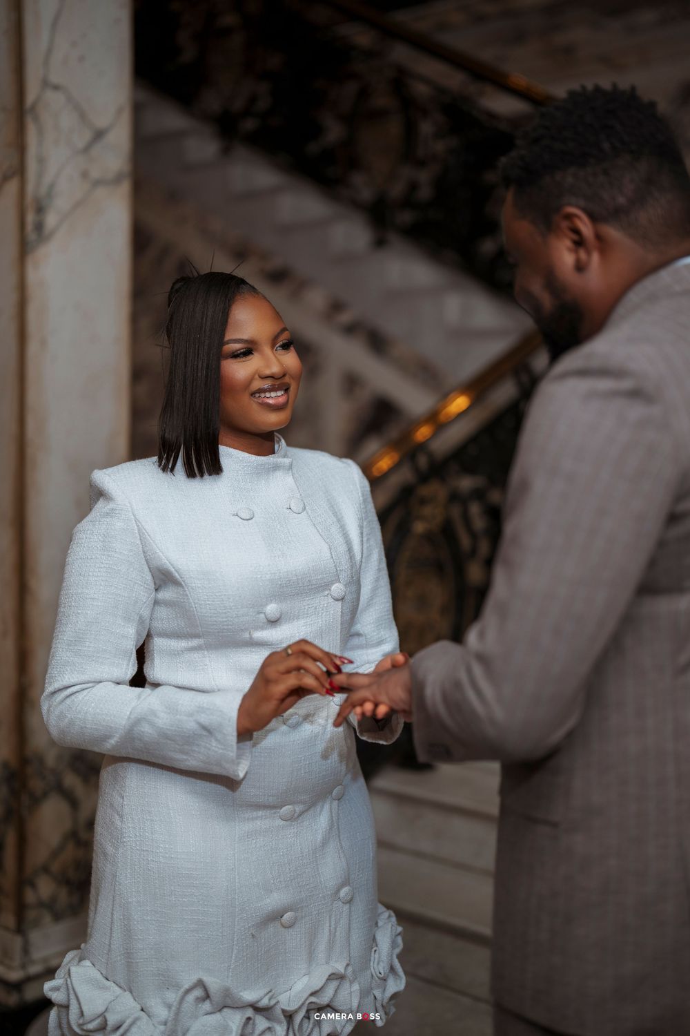 A smiling bride in a white double-breasted dress receives a ring from a groom in a gray suit during a wedding ceremony.