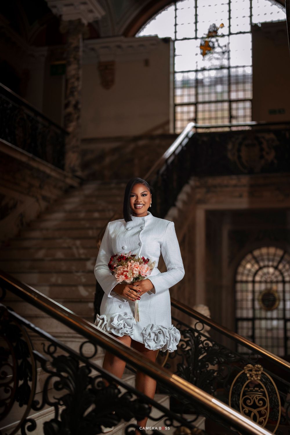 Bride in white short wedding dress holding floral bouquet on ornate staircase in elegant historic venue.