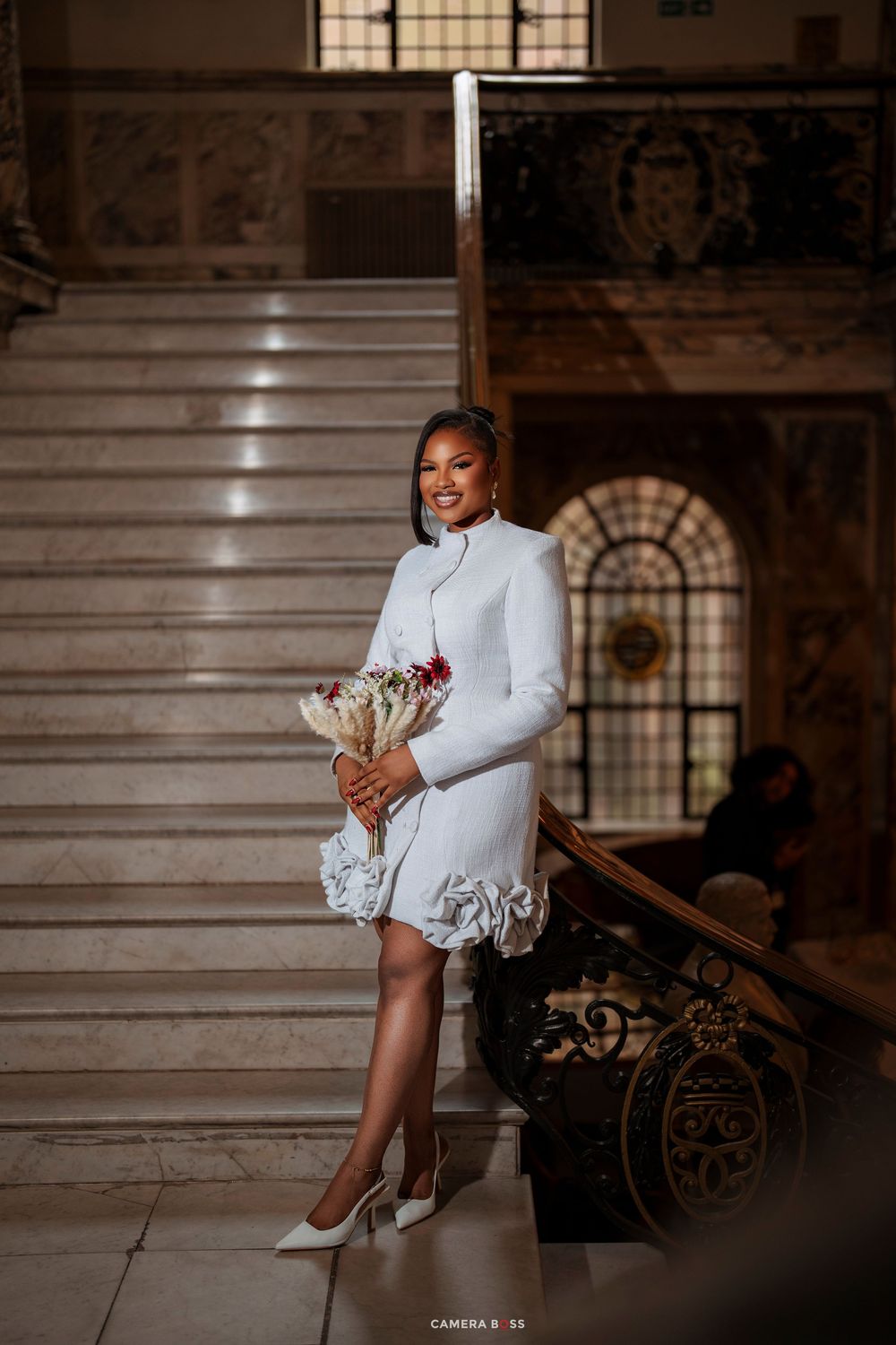 Elegant bride in white long-sleeve mini dress holding bouquet on marble staircase in ornate venue.