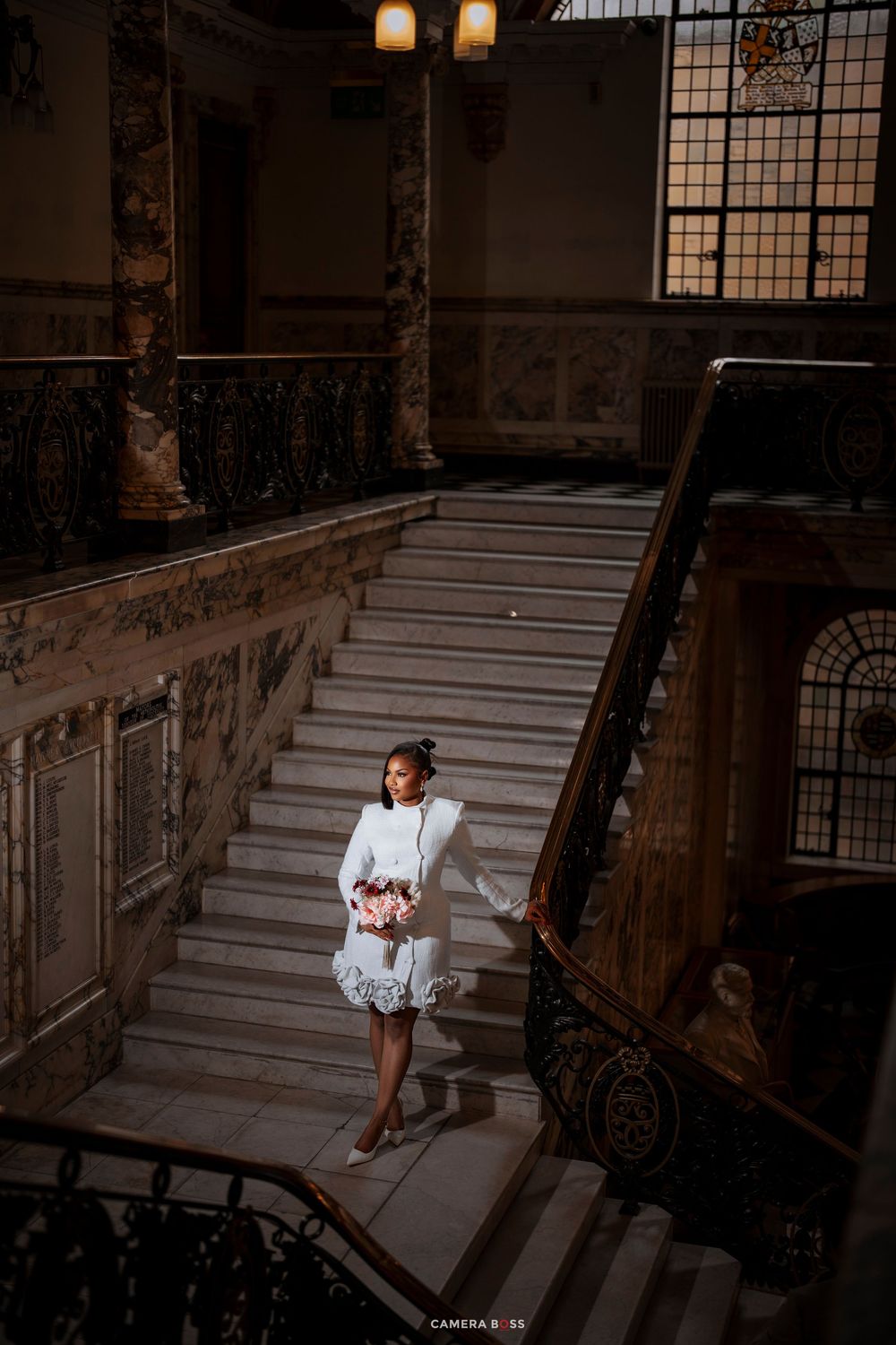 Bride in white short dress holding pink bouquet on grand marble staircase in elegant historic building.
