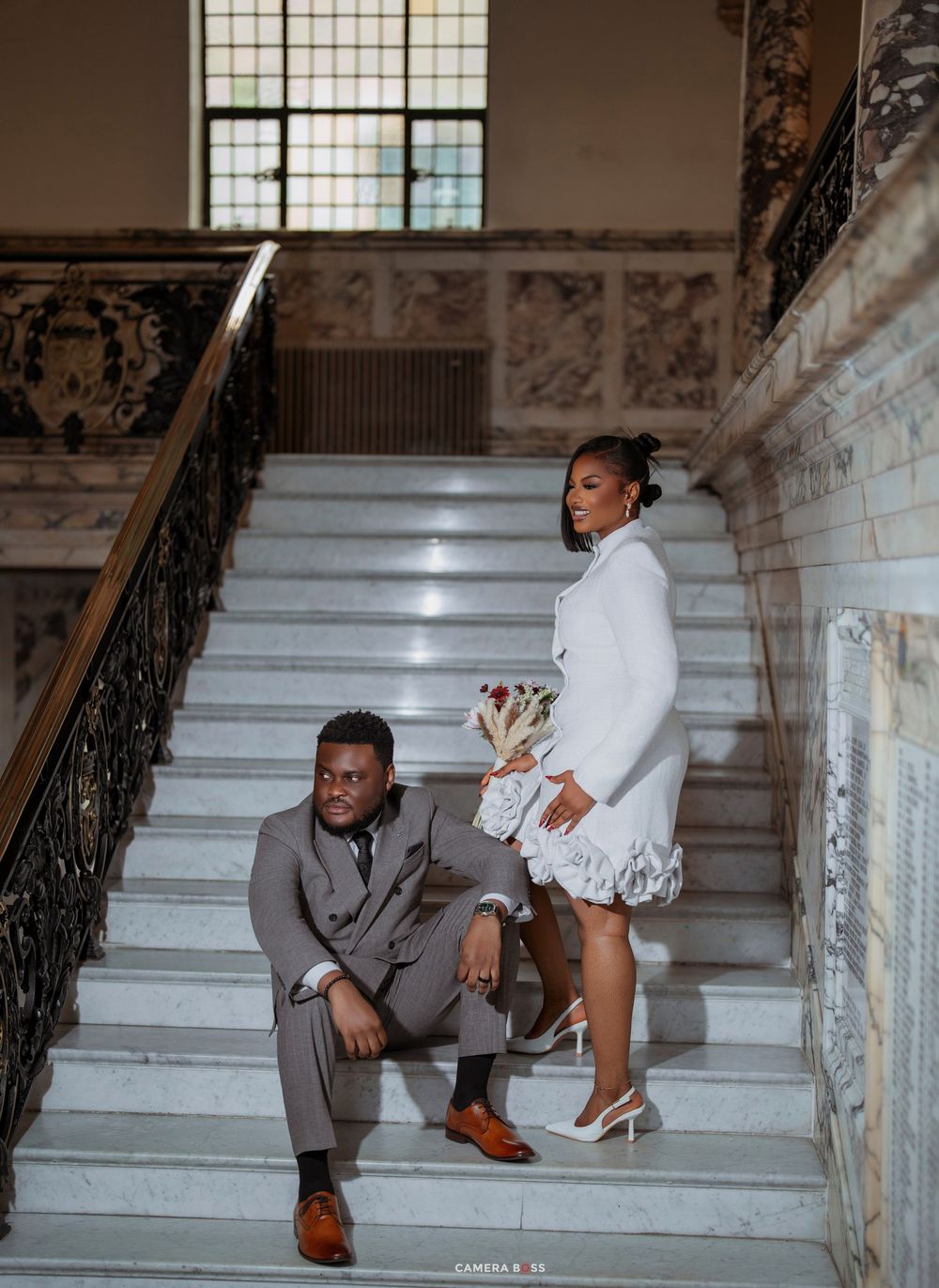 Elegant couple posing on marble staircase, man in gray suit, woman in white ruffled mini dress, grand interior.