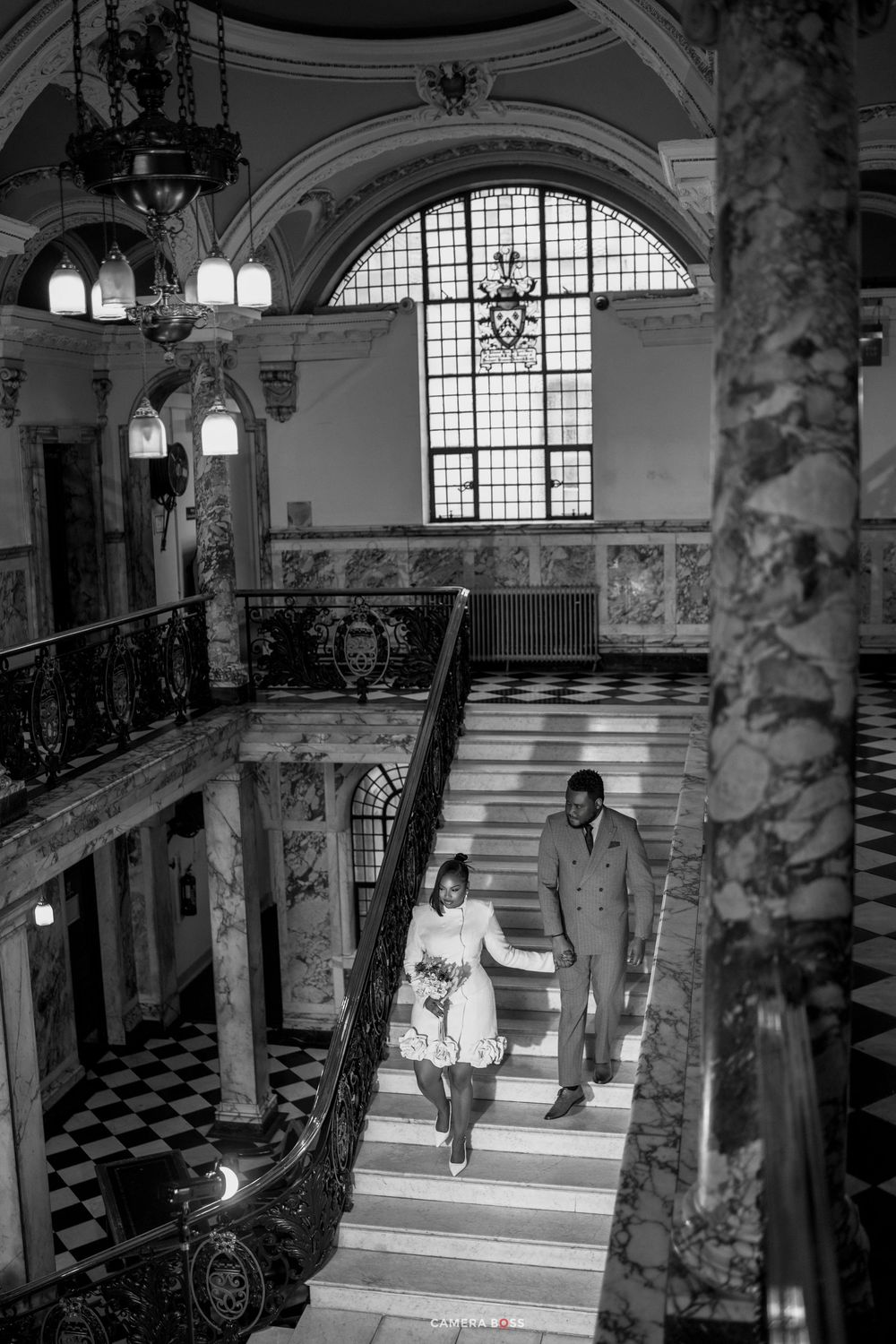 Black and white photo of a couple descending a grand marble staircase in an elegant historic building.