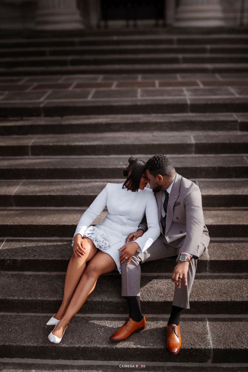 Elegant couple sharing an intimate moment on grand stone steps, woman in white dress and man in gray suit.
