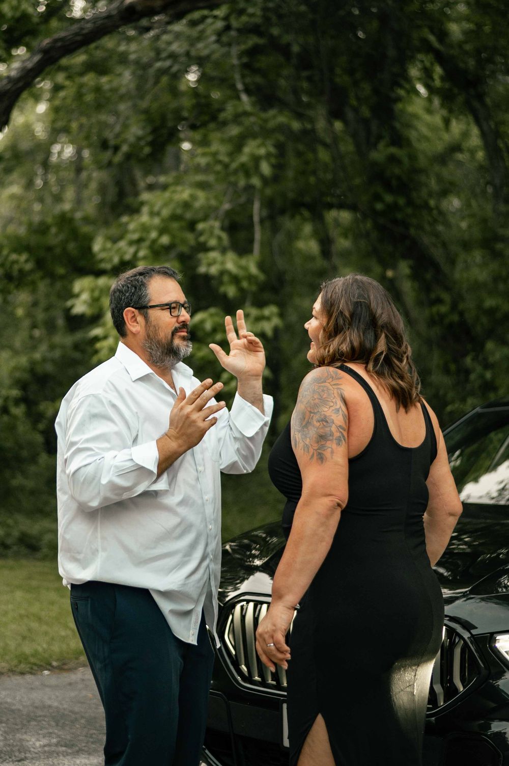 A bearded man in a white shirt gestures while talking to a tattooed woman in a black dress near a car outdoors.