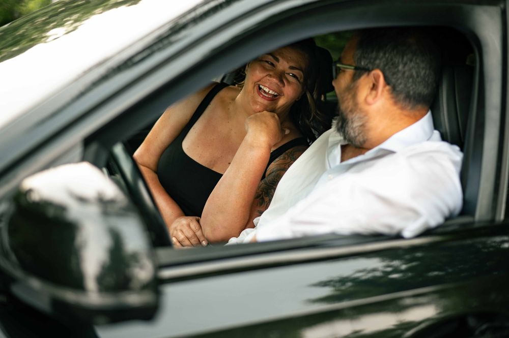 A smiling woman in a black top chats with a bearded man in a white shirt inside a car.