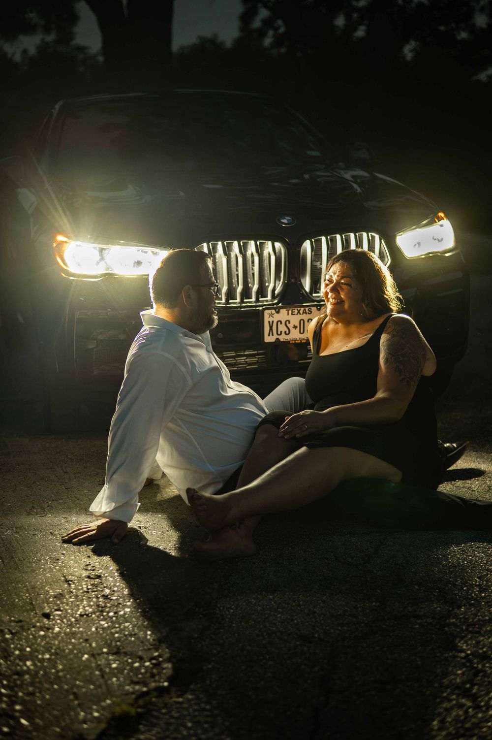 Couple sitting on pavement in front of illuminated BMW SUV headlights at night in a romantic pose.