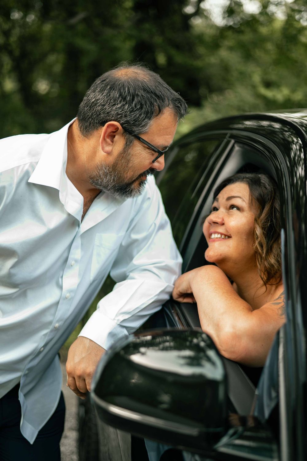 A man in a white shirt leans toward a smiling woman sitting in a car window, sharing a warm moment outdoors.