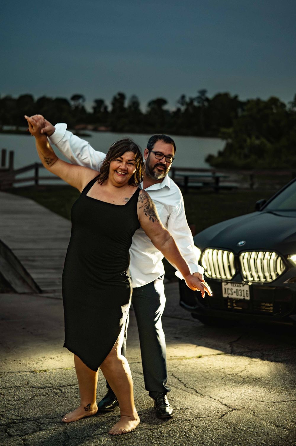 Couple dancing joyfully at dusk in front of a black BMW with glowing headlights near a waterfront.