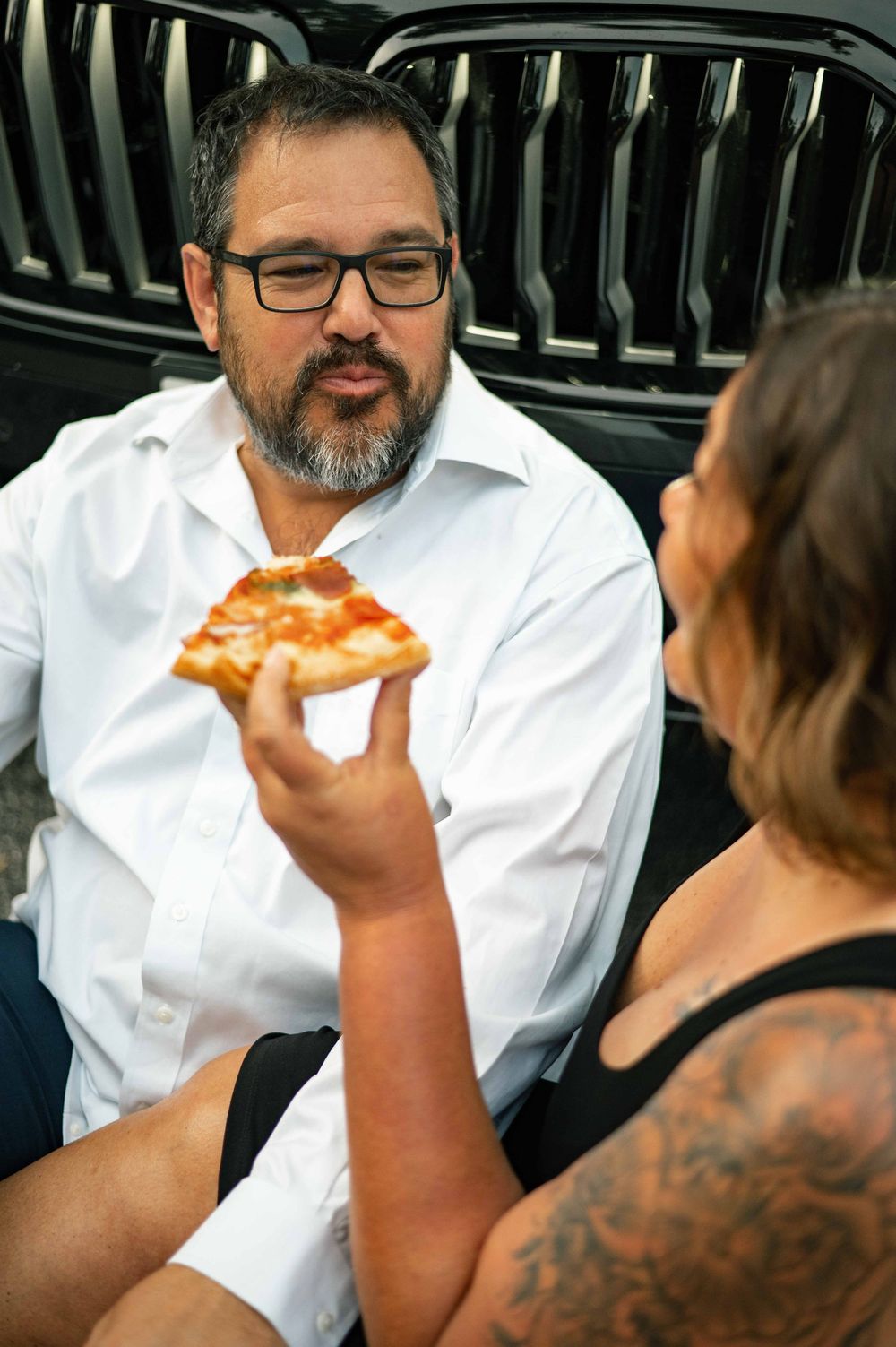 Man with glasses in white shirt being offered a pizza slice by a tattooed person near a black car.