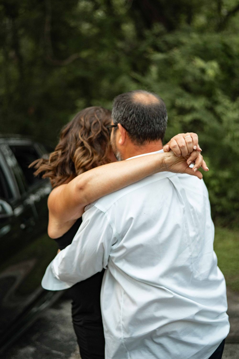 Two people share a warm embrace outdoors near a dark vehicle, surrounded by lush green trees.