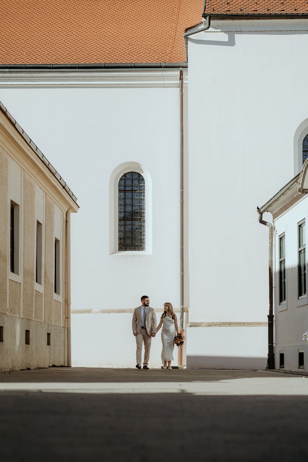 Bride and groom holding hands walking between white European buildings in sunlight.