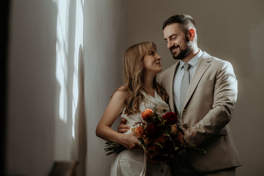 Happy couple in wedding attire holding autumn bouquet, smiling at each other in soft natural light.