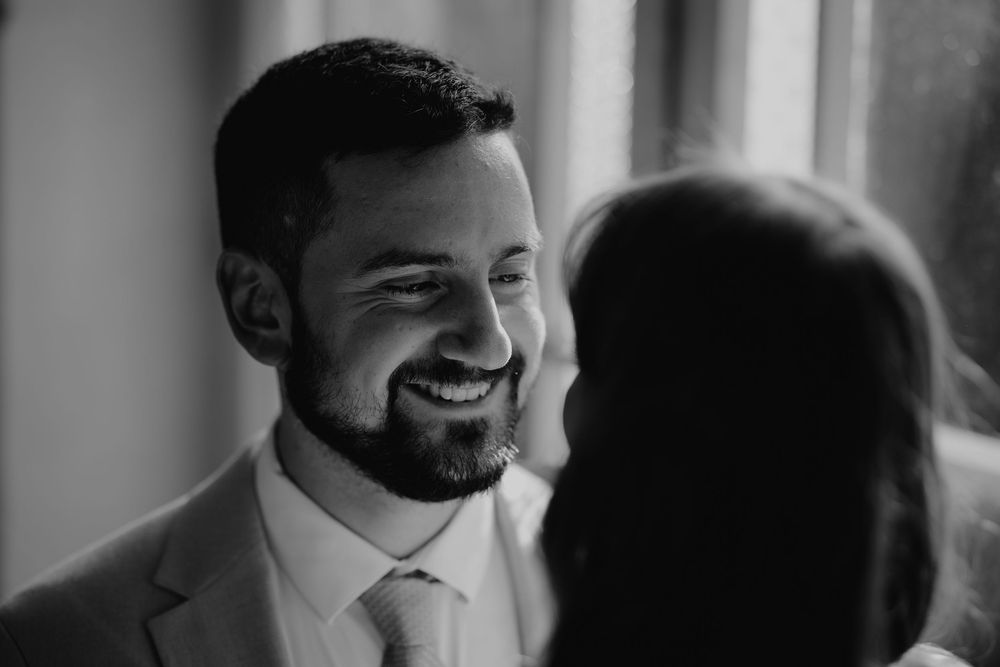 Groom smiling joyfully at bride in black and white wedding portrait near window.