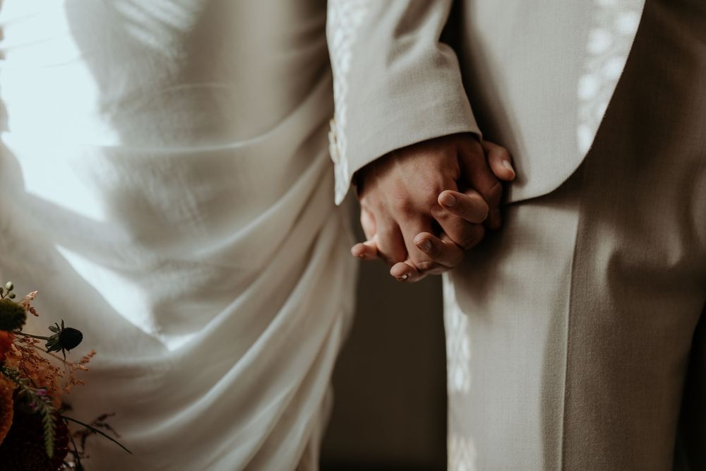Bride and groom holding hands tenderly, wearing white dress and beige suit on wedding day.