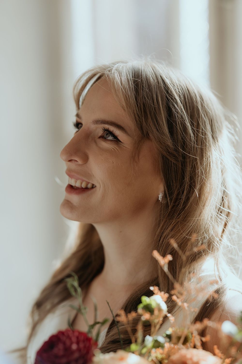 Radiant bride smiling upward holding autumn floral bouquet in soft natural window light.