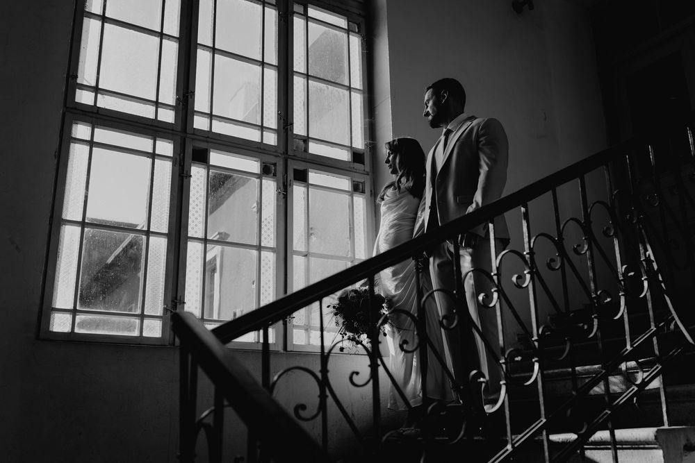 Elegant black and white photo of bride and groom standing on ornate staircase by window.