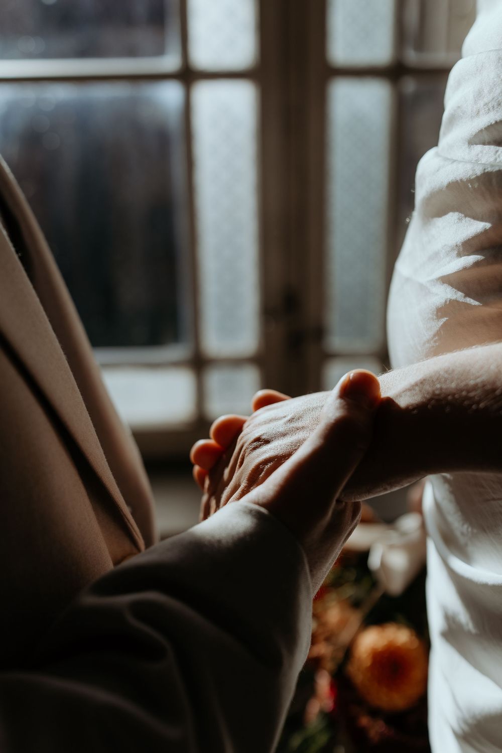 Groom gently holding brides hand near window in soft romantic wedding day lighting.
