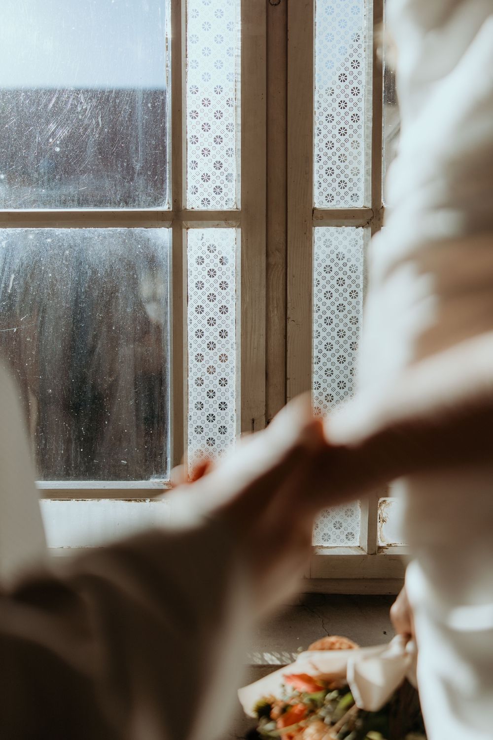 Soft light filters through a vintage lace-curtained window illuminating delicate white fabric and flowers below.