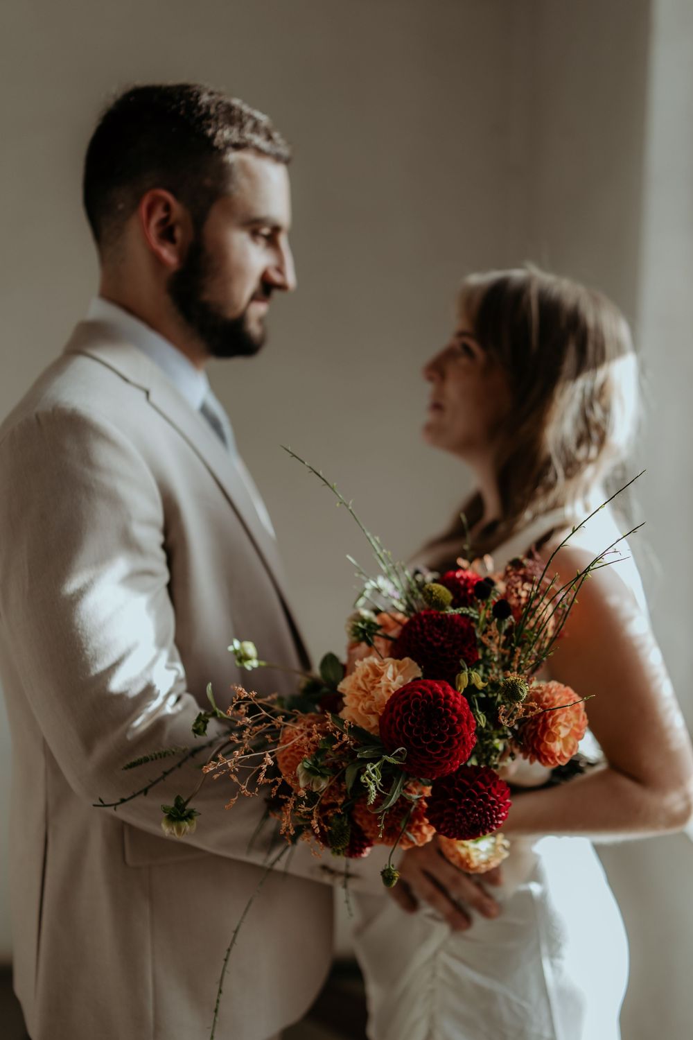 Bride and groom share an intimate moment holding a vibrant autumn bouquet of red and orange dahlias on their wedding day.