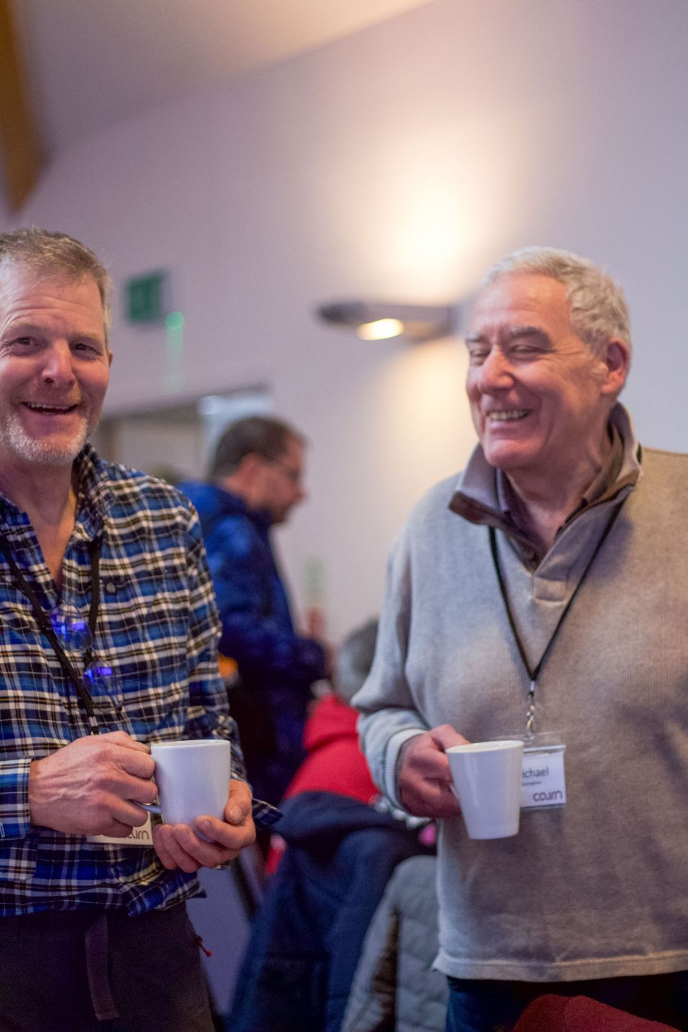Two smiling middle-aged men holding coffee mugs and chatting at an indoor networking or community event.