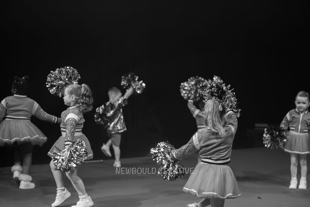 Young cheerleaders in uniform performing with pom-poms on stage in black and white photo.
