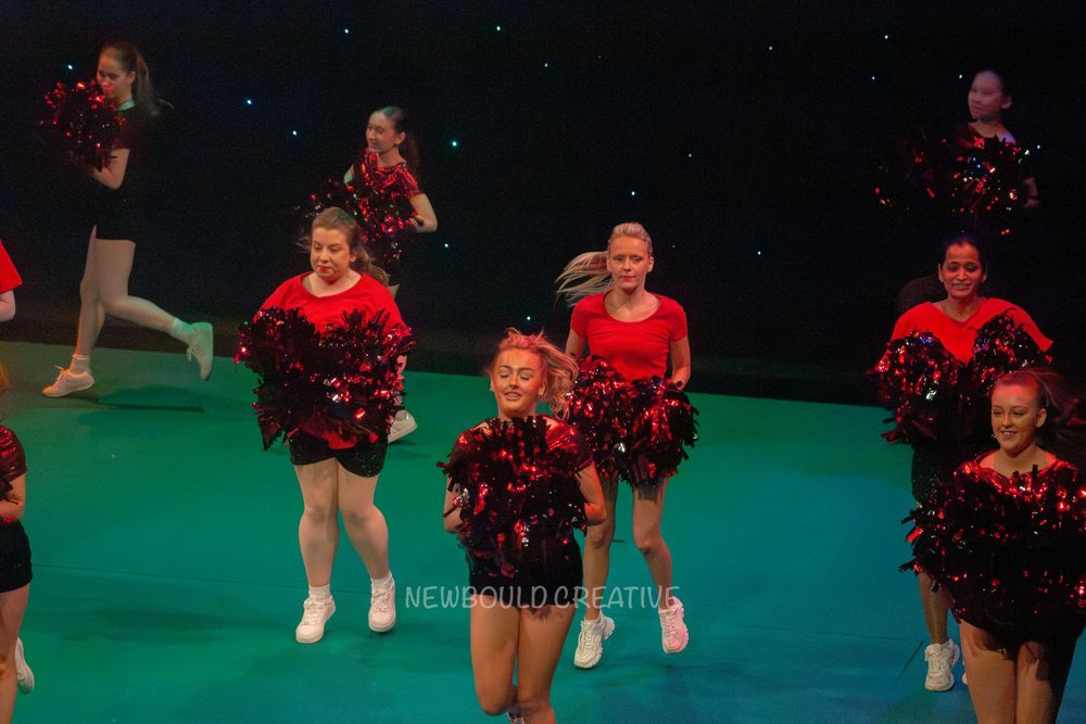 Young cheerleaders in red and black uniforms performing with pom-poms on a green stage.