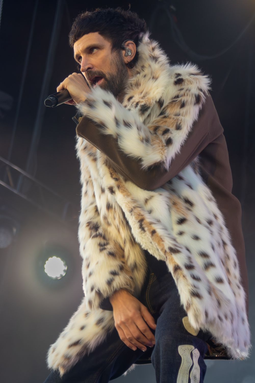 Male singer performing on stage in a white leopard print fur coat, leaning forward holding a microphone.