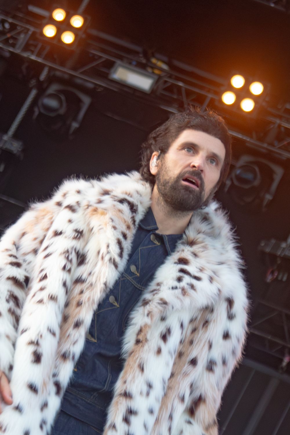 Male performer on stage wearing a striking white spotted fur coat over denim shirt under bright stage lights.