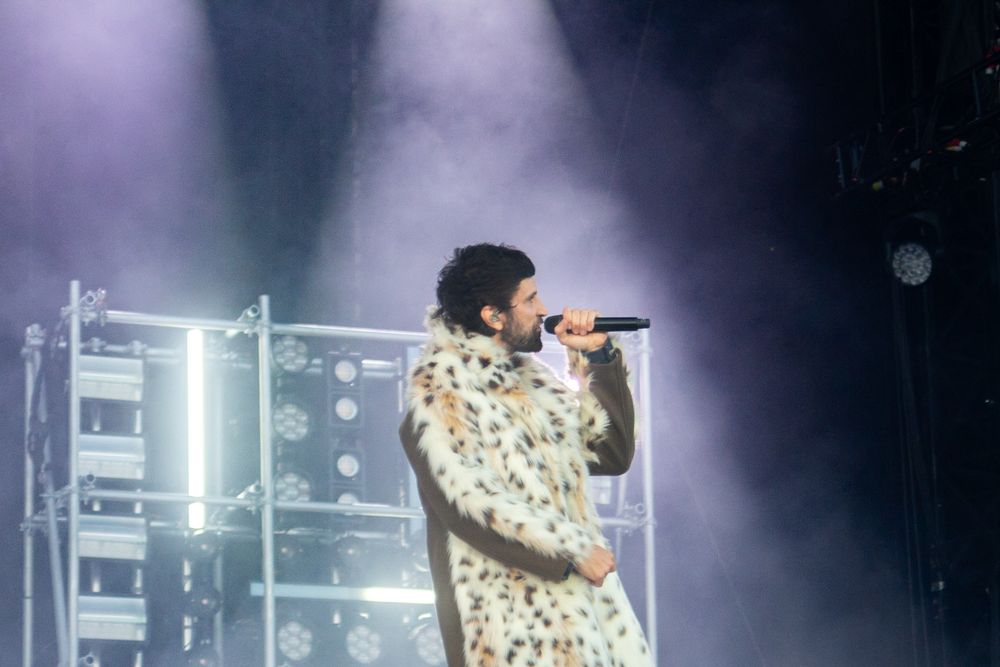 Male artist performing on stage in a spotted fur coat with dramatic fog and light display behind him.