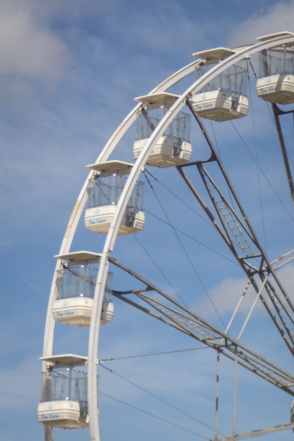 White ferris wheel gondolas against a blue cloudy sky at an outdoor festival or fairground.
