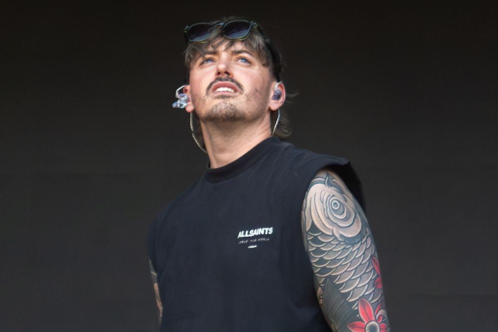 Tattooed male performer on stage wearing a black sleeveless shirt with sunglasses resting on his head.